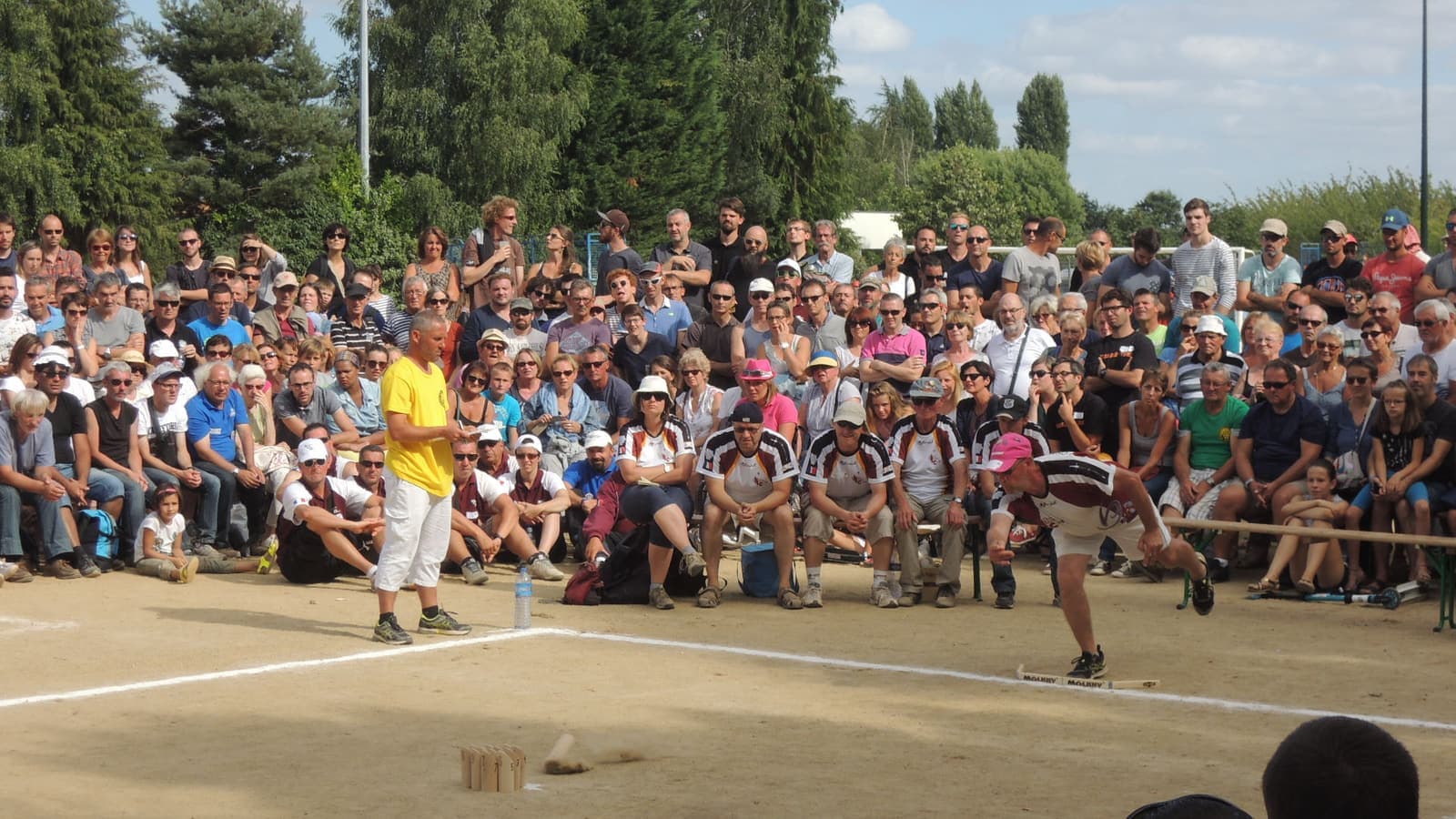 Ein Spieler hat das Mölkky gerade unterhand losgelassen; das Wurfholz fliegt über dem Sand, am anderen Ende stehen die zwölf Kegel, Publikum schaut zu.