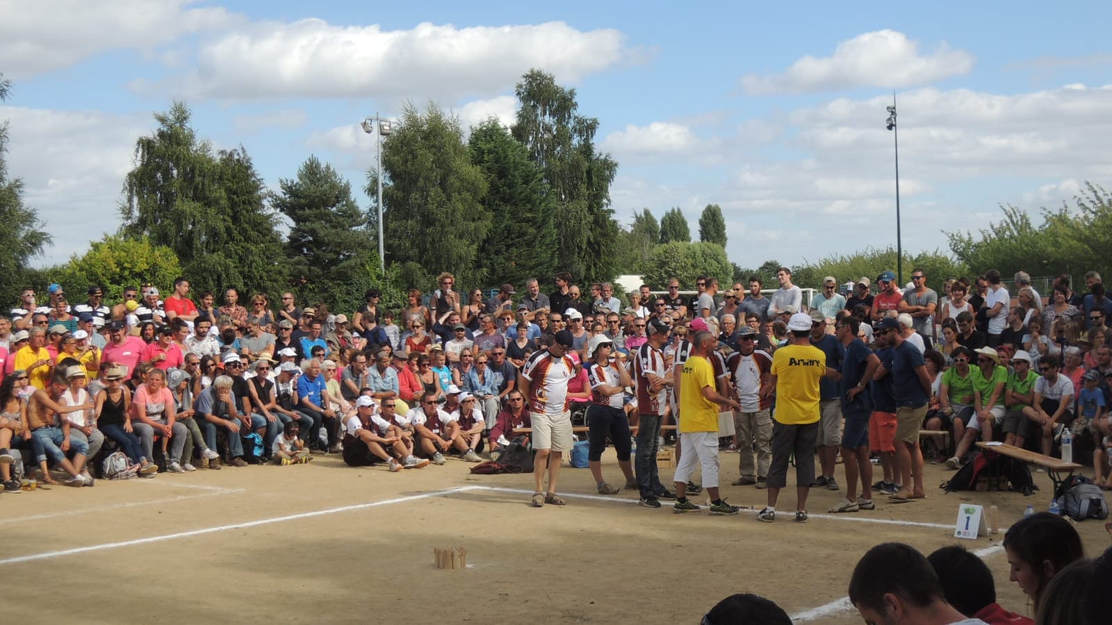 Eine große Menschenmenge steht und sitzt rund um einen Sandplatz bei einem Mölkky-Turnier und verfolgt zwei Teams und einen Schiedsrichter an der Wurflinie.