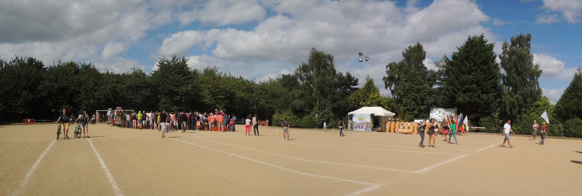 Een breed zandveld met uitgezette banen op een Mölkky-toernooi, met spelers en toeschouwers langs één zijde.